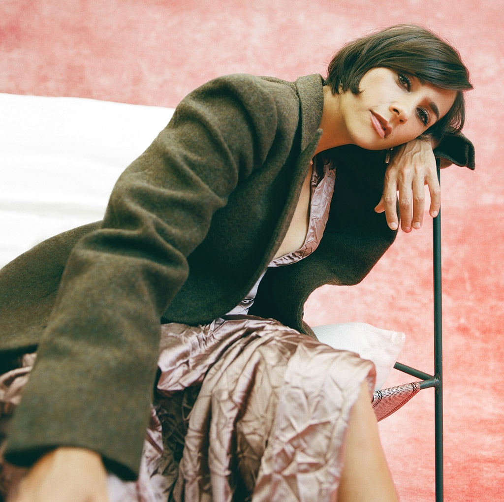 Woman in gray suit leaning on chair arm against pink backdrop