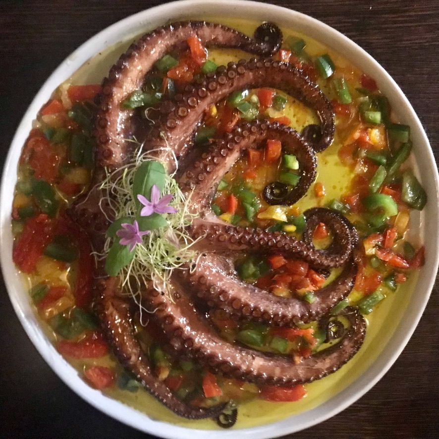 Bowl of octopus tentacles in yellow sauce with peppers, sprouts, and small purple flowers