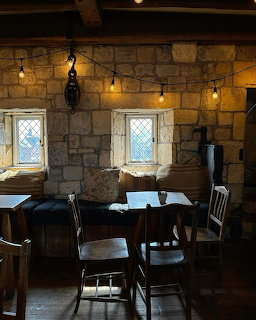 Cozy stone-walled cafe interior with wooden beams, string lights, small windows, and dark wooden tables and chairs