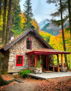 Stone cottage with red window frames and porch surrounded by colorful autumn trees in a forest