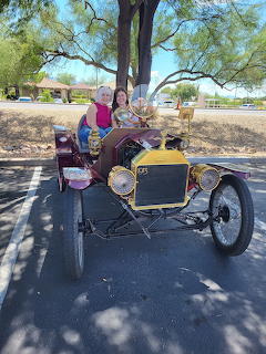 Two people sitting in a vintage 1913 Model T Ford Speedster parked under a tree on a sunny day