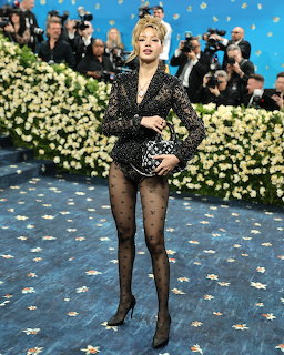 Woman in black sequined dress and tights posing on blue carpet with white flowers