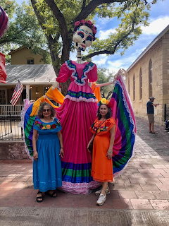Two women posing with a large Dia de los Muertos catrina skeleton figure in a colorful dress outdoors