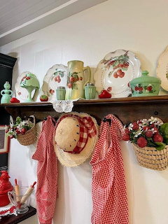 Vintage kitchen shelf with strawberry plates, green jadeite containers, red gingham towels, and a straw hat