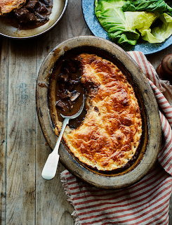Oval baking dish with golden cheese-topped pie, spoon, braised meat and lettuce on wooden table