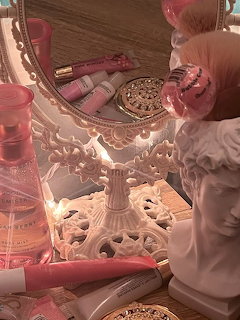 Vanity table with ornate white mirror, porcelain bust, pink perfume bottles, lipsticks, and gold compact