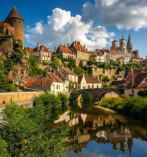 Medieval French town with towers and stone buildings reflected in a calm river surrounded by greenery