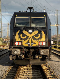 Black electric locomotive with yellow owl design on front, red lights, on train tracks