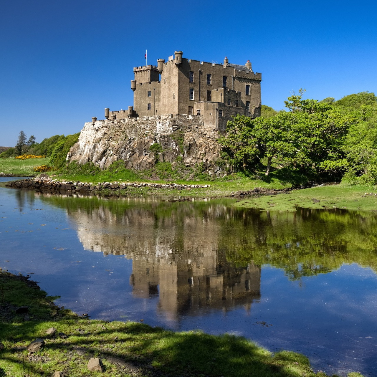 Stone castle on a rocky outcrop reflected in calm water with trees and blue sky