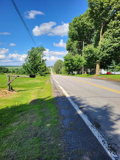 Country road with yellow center line bordered by green trees and grass under blue sky with clouds