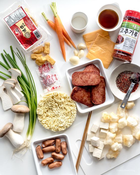 Overhead view of Korean army stew ingredients including spam, sausages, noodles, tofu, and vegetables on white background