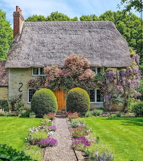 Stone cottage with thatched roof, rounded topiary, flower garden and gravel path leading to wooden door
