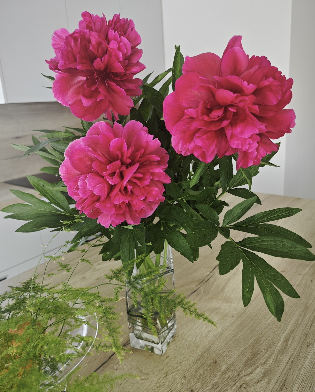 Bright pink peonies in a clear glass vase on a wooden table with green leaves