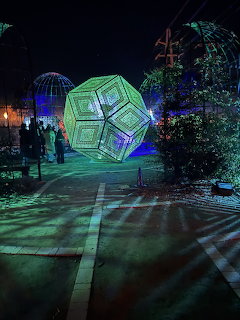 Large glowing green geometric light sculpture in a garden at night with visitors walking nearby