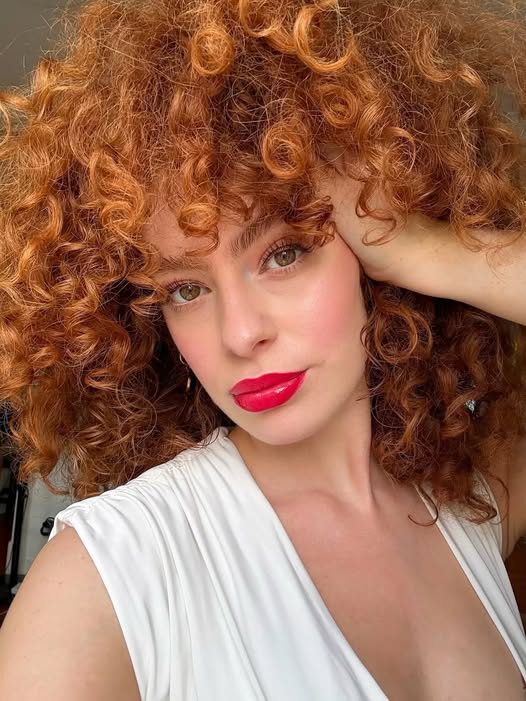 Young woman with curly ginger hair and red lipstick posing for a selfie in a white top