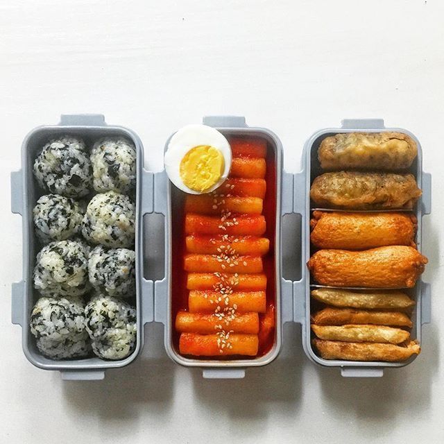 Three lunch boxes with rice balls, carrot sticks and dip, and assorted bread rolls on a white surface