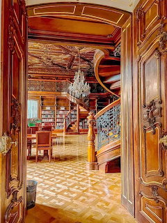 Ornate library interior with carved wood, staircase, chandelier, and bookshelves