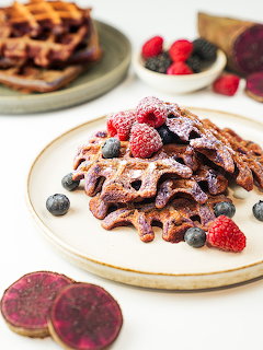 Stack of purple waffles topped with raspberries and blueberries on a white plate with purple sweet potato slices