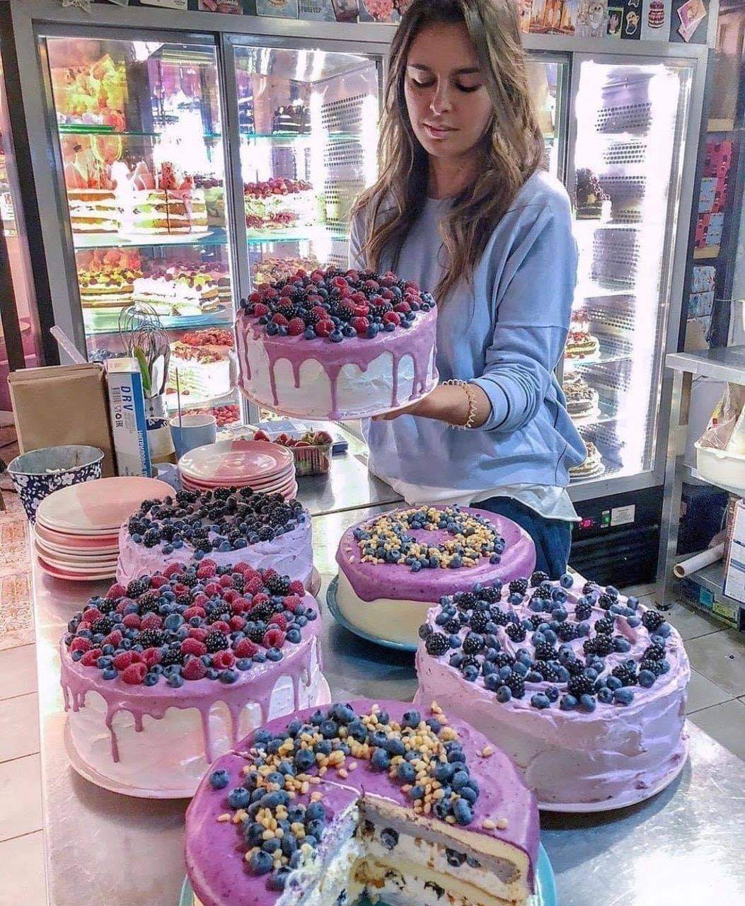 Woman holding berry-topped cake in bakery with multiple decorated cakes on counter