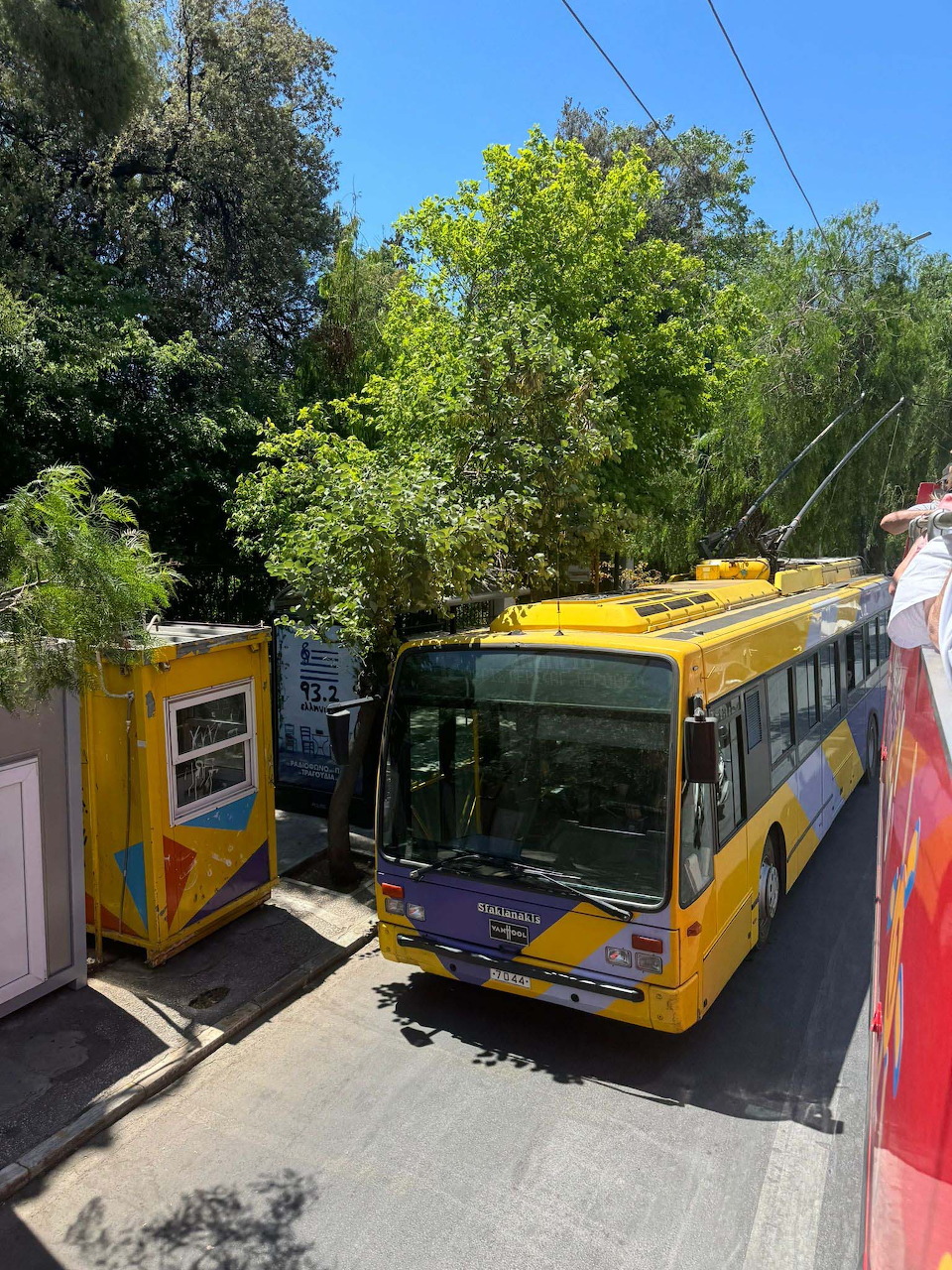 Yellow Athens trolleybus passing a ticket kiosk on a tree-lined street on a sunny day
