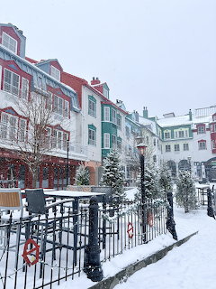 Snow-covered village square with colorful multi-story buildings, evergreen trees, and iron fence in winter