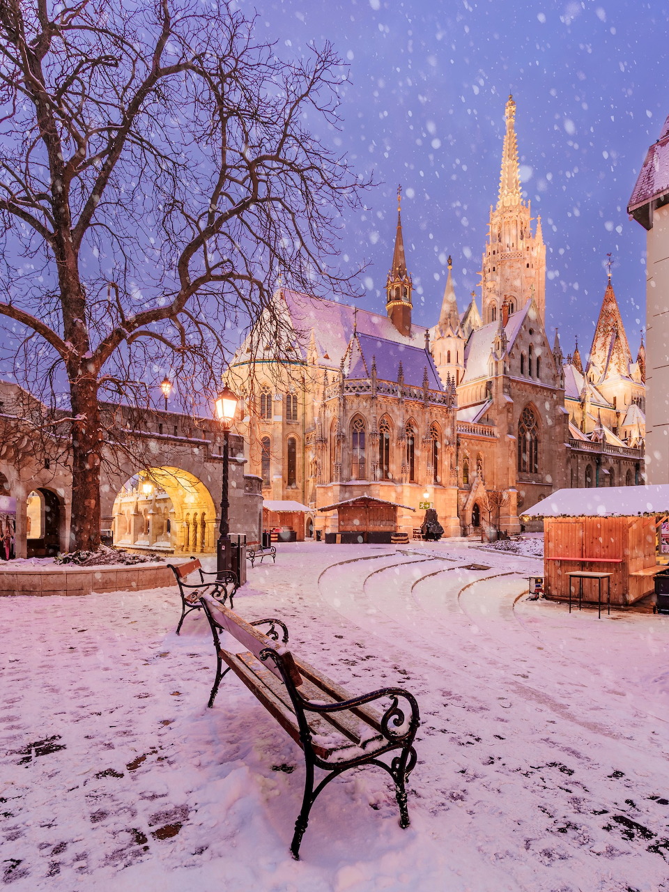 Matthias Church in Budapest on a snowy evening with warm lights, bare trees, and a bench