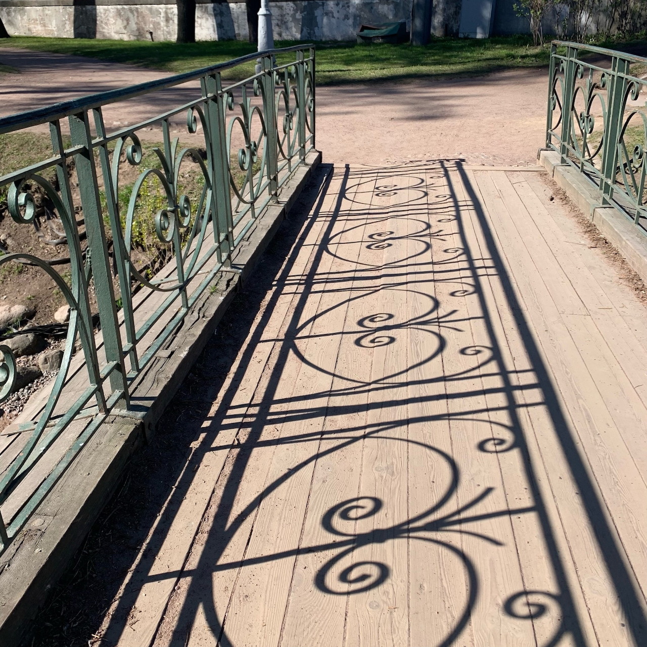 Ornate metal fence casting curly shadows across a wooden bridge walkway