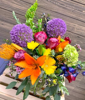 Bright mixed flower bouquet with orange lilies, purple alliums, roses, and assorted blooms in vase on wood surface