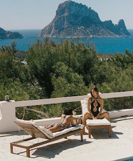 Two women in black swimwear on sun loungers on a white terrace with Mediterranean sea and rocky island view