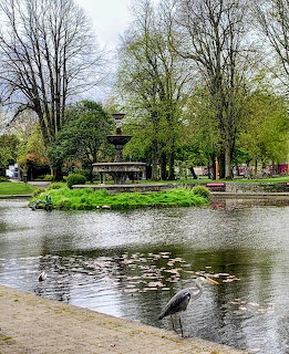 Fountain in a park pond with trees and a heron on the shore
