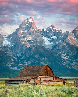 Rustic wooden barn in green field with Grand Teton mountain peaks and pink clouds at sunrise
