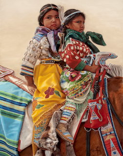 Two young Native American girls in traditional dress sitting on a horse with colorful blankets and beadwork