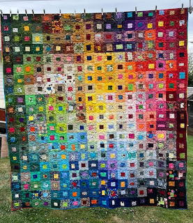 Handmade patchwork quilt displaying rainbow gradient of colorful fabric squares hung outdoors