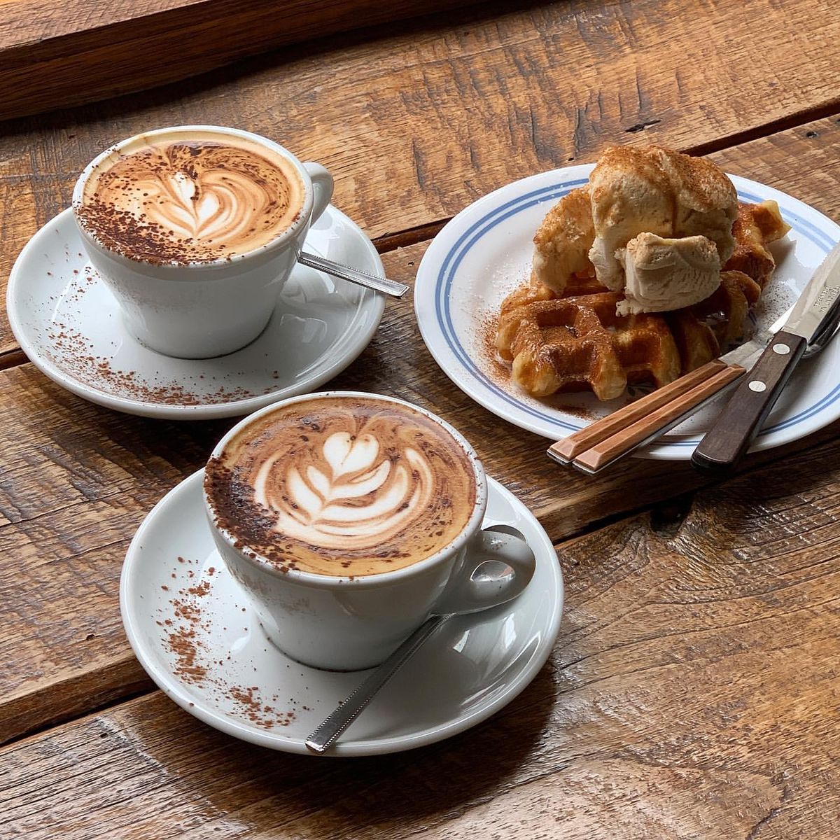 Two cappuccinos with foam art and a Belgian waffle with butter on white plates over wooden table