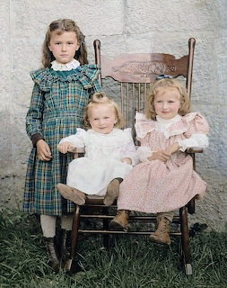 Colorized vintage photo of three little girls in Victorian dresses posing with a wooden rocking chair
