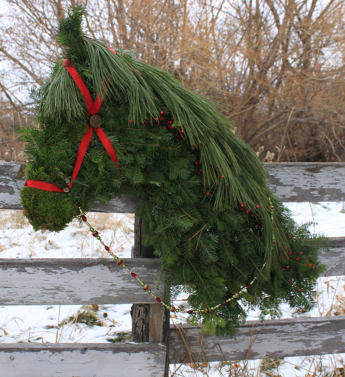 Horse head shaped wreath made of evergreen branches with red ribbon on white fence in snow