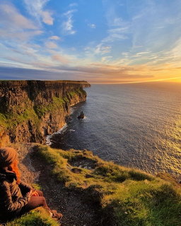 Person sitting on the edge of the Cliffs of Moher overlooking the ocean at sunset