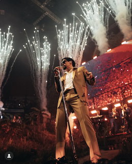 Singer in a tan suit performing on an arena stage with fireworks and a large crowd behind him