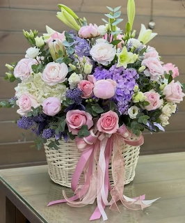 Wicker basket filled with pink roses, purple flowers, white lilies and pastel blooms with pink ribbons