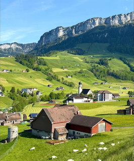 Alpine village with green hills, farm buildings, sheep, and a rocky mountain ridge beneath blue sky