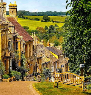 Scenic view down a traditional English village street with honey-colored stone houses and green countryside beyond