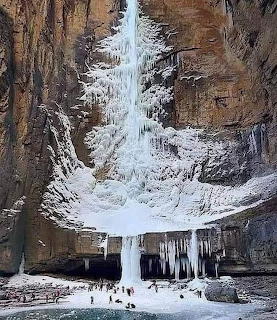 Tall frozen waterfall with icicles hanging from rocky cliff above turquoise pool