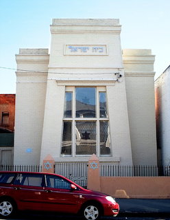 White Art Deco synagogue building with tall arched window and Hebrew inscription above entrance