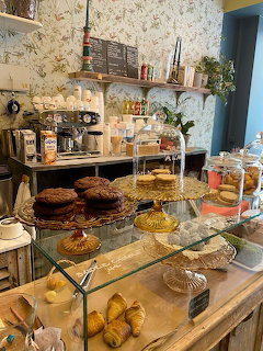 Café display counter with tiered stands holding cookies and croissants, floral wallpaper, espresso machine in background