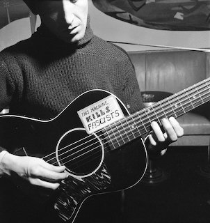 Black and white photo of man in turtleneck playing acoustic guitar with 'This Machine Kills Fascists' sticker