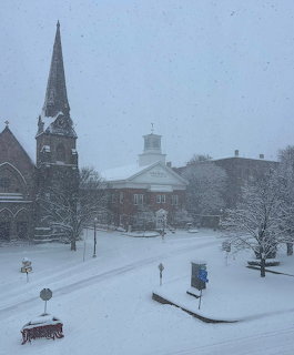 Snow-covered town center with church steeple and colonial buildings during heavy snowfall