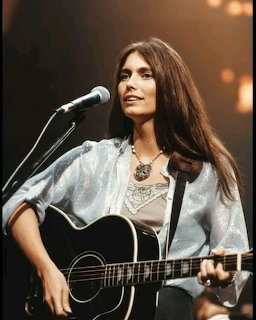 Woman with long dark hair singing into a microphone and playing acoustic guitar on stage