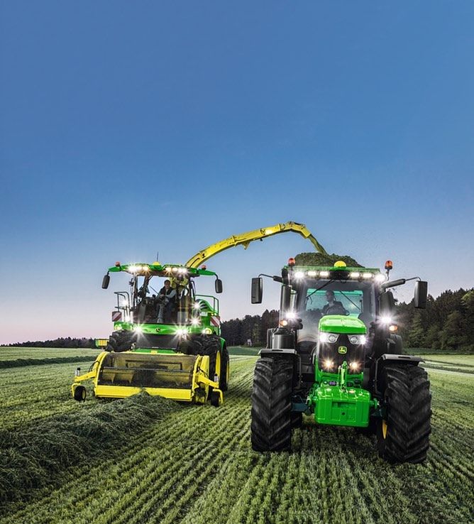 Two green tractors with bright lights harvesting a green crop field at dusk