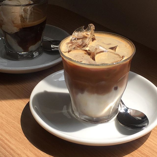Flat white coffee in a glass on a white saucer with spoon, next to another coffee on a wooden table