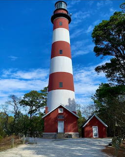 Tall red and white striped lighthouse with brick buildings at base, trees and blue sky